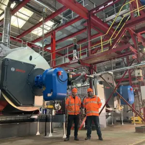 Two workers in orange safety gear stand in a large industrial facility, surrounded by red steel structures and Bosch machinery.