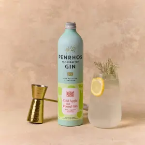 A bottle of Penrhos handcrafted gin beside a jigger and a glass of gin with a lemon slice and a sprig of herb