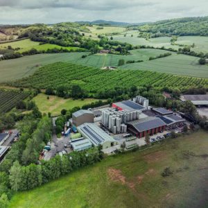 An aerial view of the Bevisol processing facility in Ledbury, Herefordshire, showcasing industrial storage tanks, solar-panelled roofing, and surrounding agricultural farmland