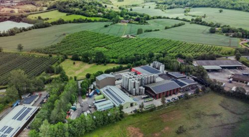 An aerial view of the Bevisol processing facility in Ledbury, Herefordshire, showcasing industrial storage tanks, solar-panelled roofing, and surrounding agricultural farmland