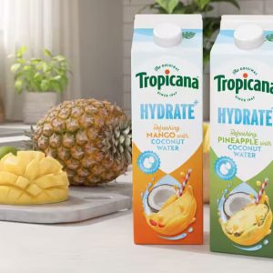 Two cartons of Tropicana Hydrate plus coconut water drink in Mango and Pineapple flavors on a bright kitchen counter next to a fresh whole pineapple, sliced mango, and coconut